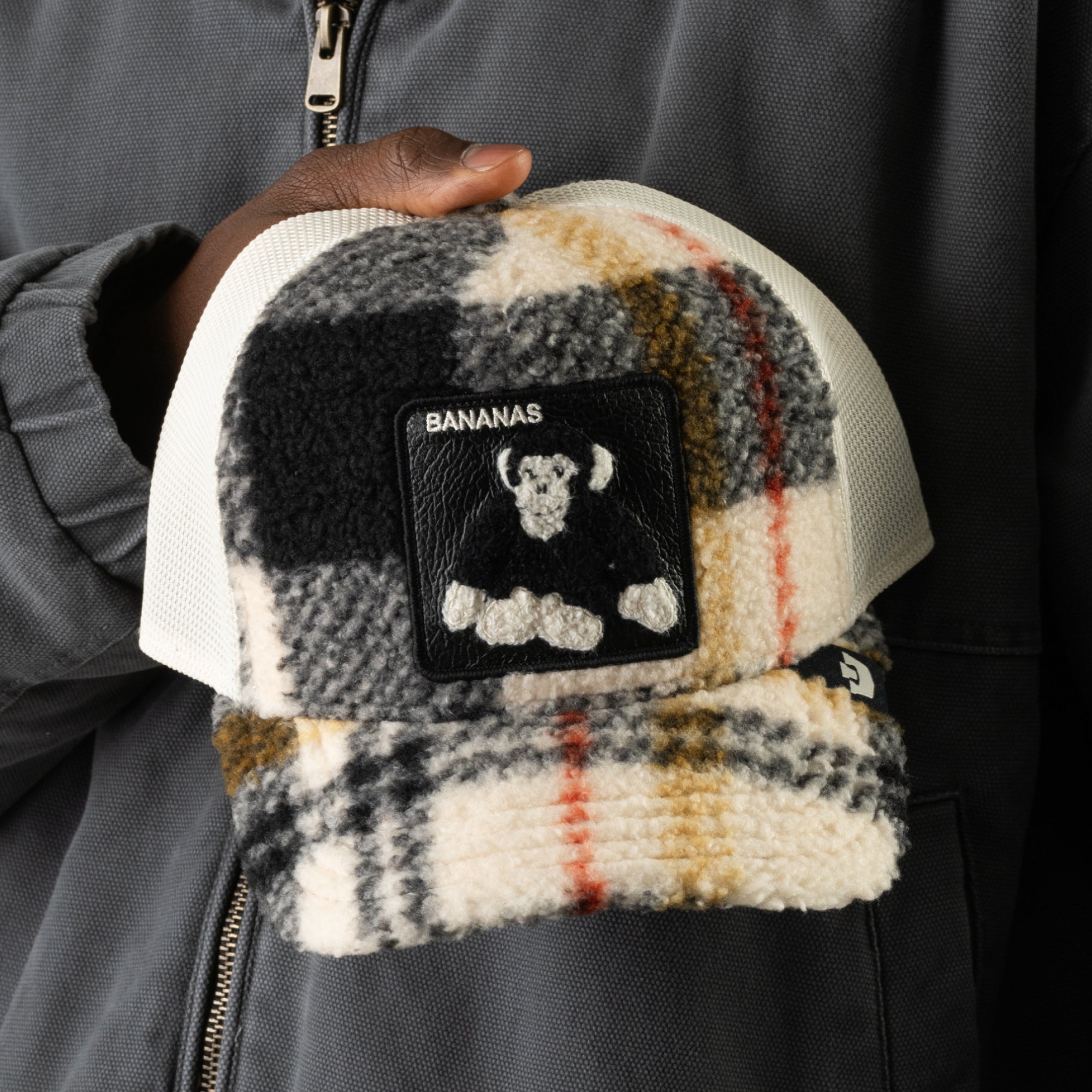 A person in a gray jacket holds the Goorin Bros. Double Stuffed - Bananas hat, a playful plaid fleece trucker hat featuring a patch with “BANANAS” and a cartoon monkey, complete with an adjustable snapback for the perfect fit.