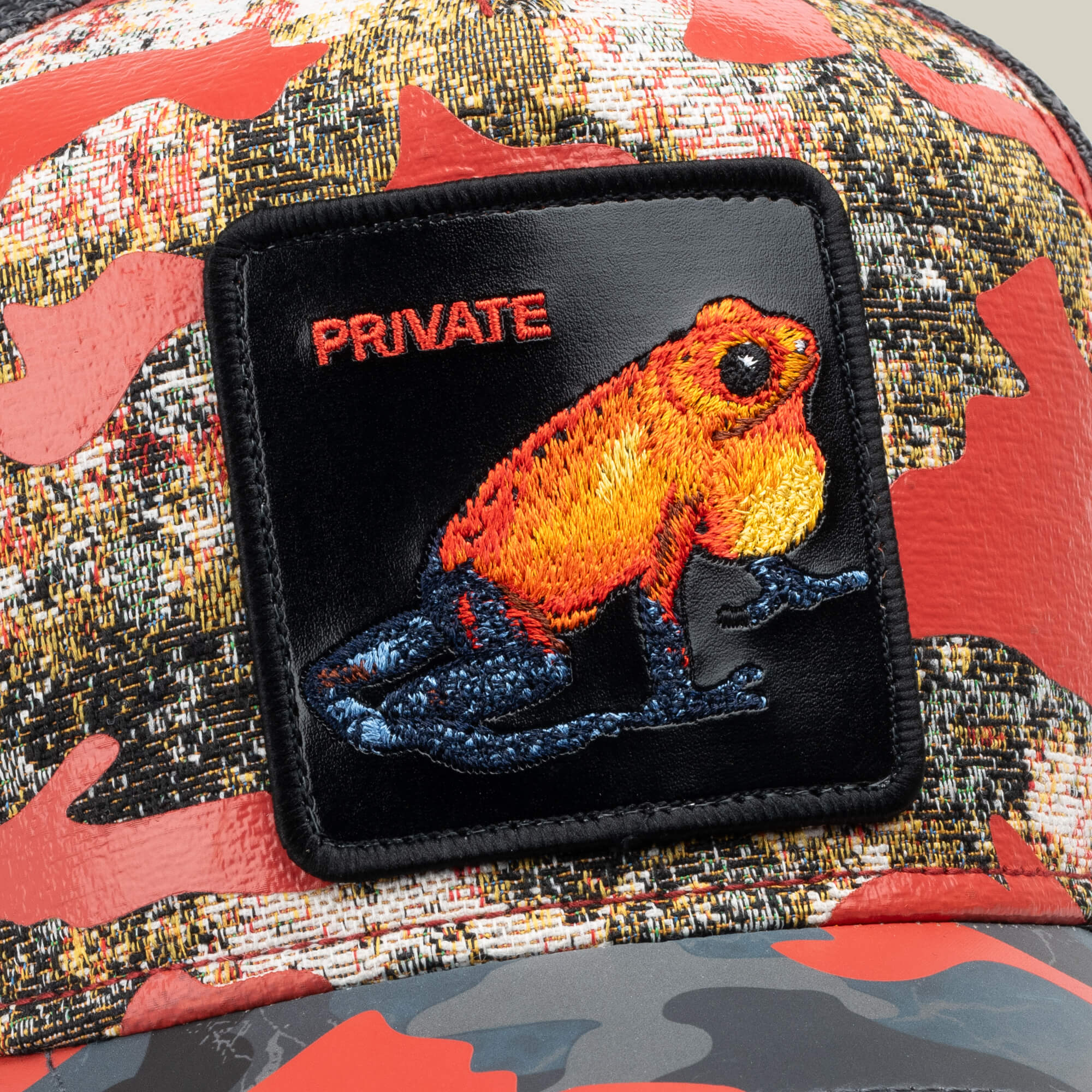 Close-up of the Goorin Bros. "Secret Service" red camo trucker hat, featuring a patch with an orange frog and the word "PRIVATE" embroidered in red.