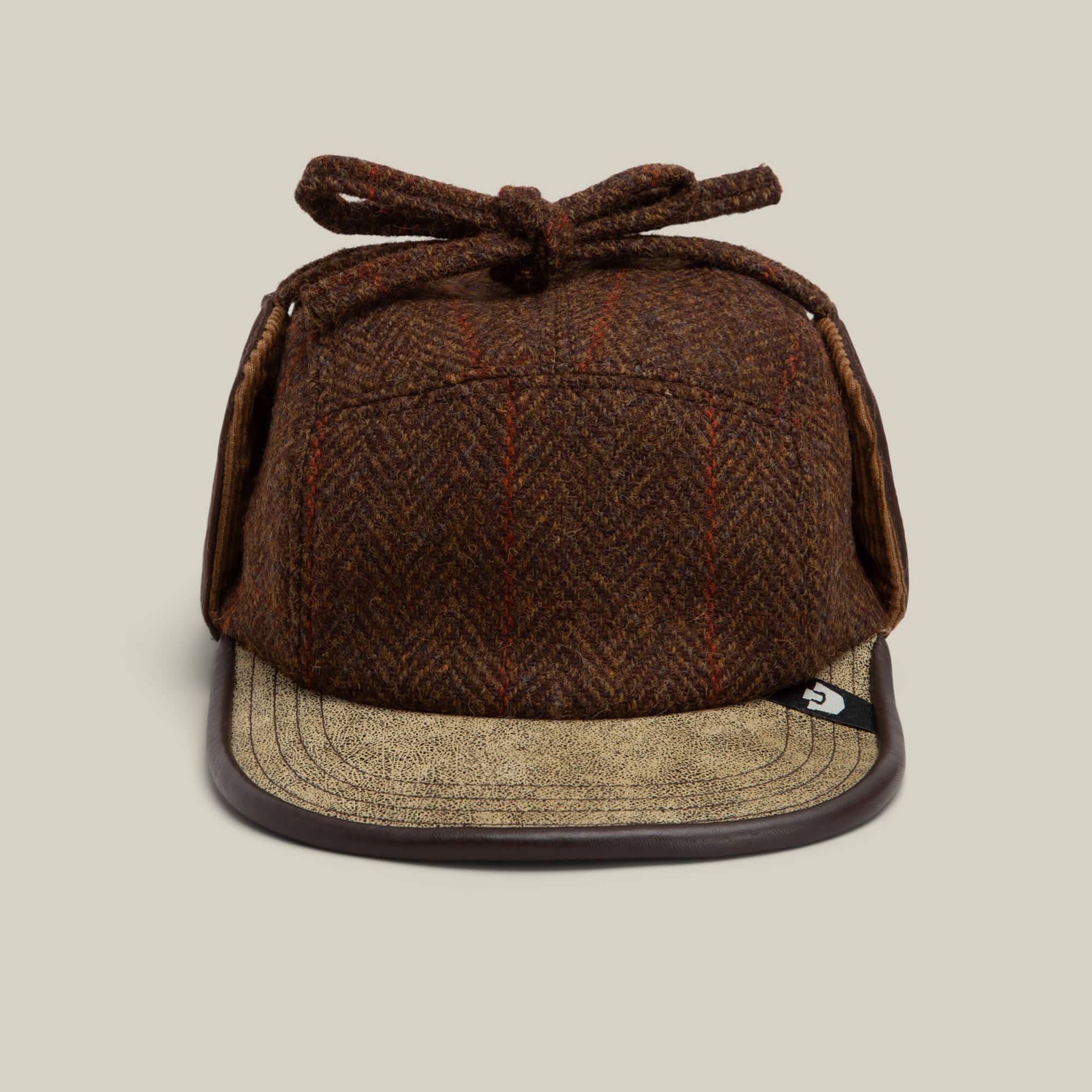 The Goorin Bros. Frazier Lake cap in AMBER features a tweed wool finish, ear flaps, a top bow, and a tan curved leather brim, photographed against a neutral background.