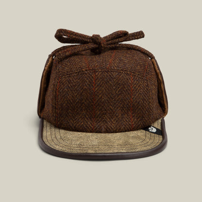 The Goorin Bros. Frazier Lake cap in AMBER features a tweed wool finish, ear flaps, a top bow, and a tan curved leather brim, photographed against a neutral background.