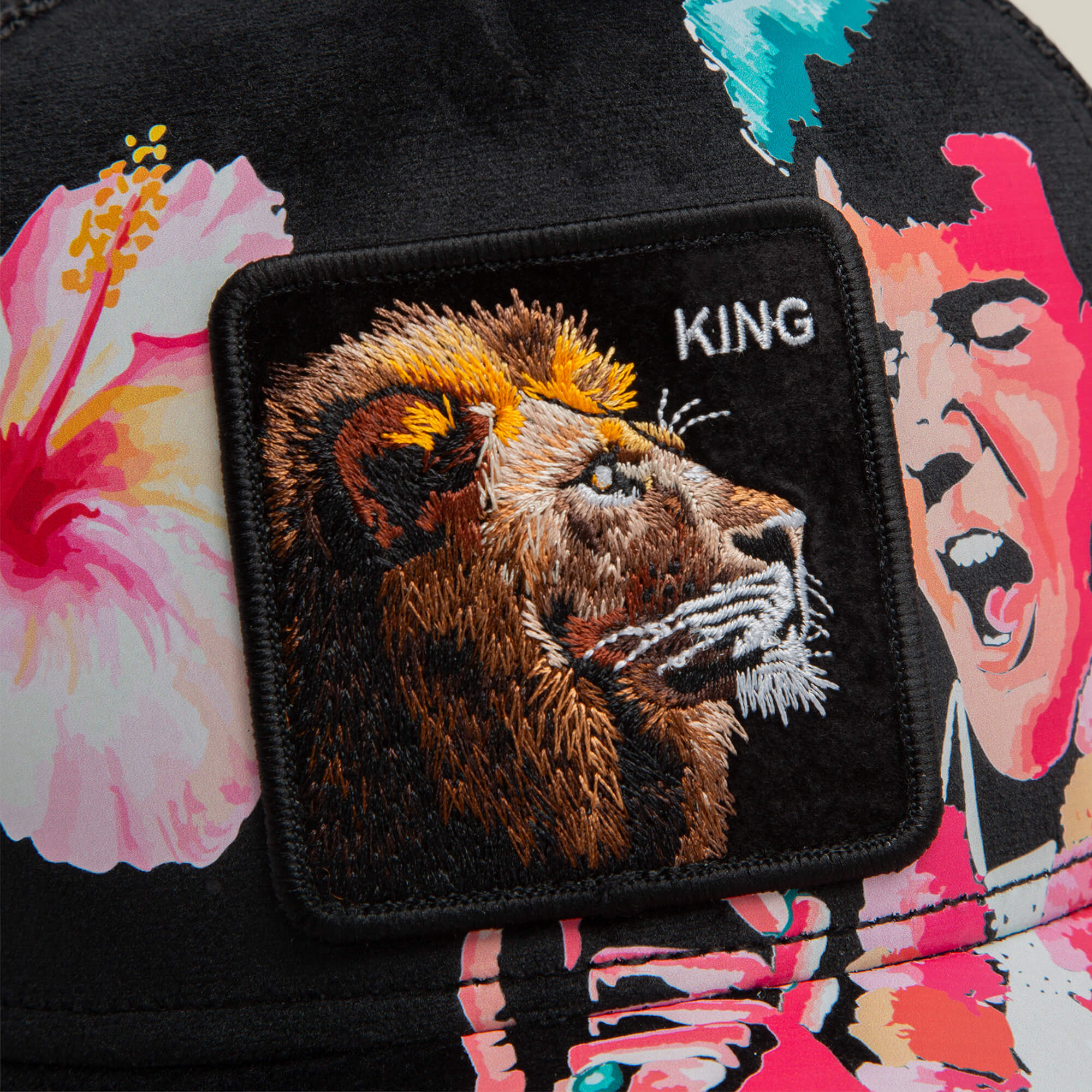 The Goorin Bros. Uh Huh King is a black hat with a colorful floral pattern, a blue suede underbrim, and features an embroidered lion patch labeled UH HUH KING, with part of an illustrated face in the background.