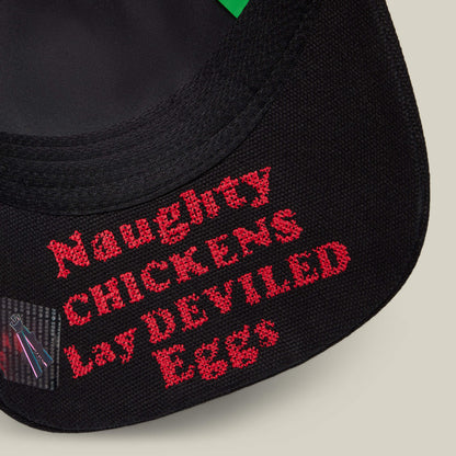 Close-up of black fabric with red embroidered text, "Naughty CHICKENS Lay DEVILED Eggs," from Goorin Bros.’ Middle of No Where collection—ideal for fans of home cooking or fried chicken.