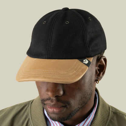 A man wearing the Goorin Bros. Bixby hand-sewn wool and leather cap in black and beige, paired with a green jacket and striped shirt, looks slightly down against a plain background.