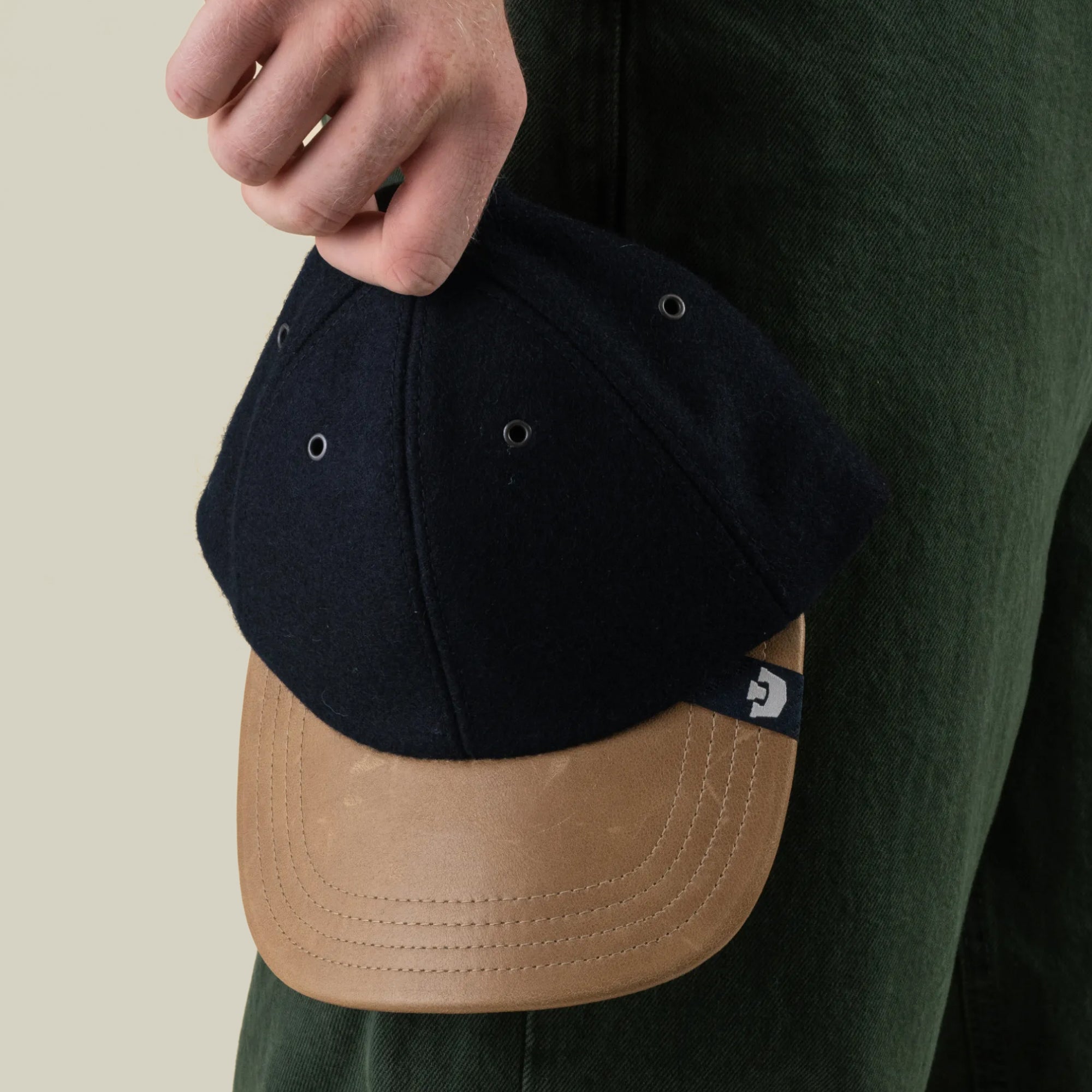 A person holding the Goorin Bros. Bixby—an expertly hand-sewn black and tan wool leather baseball cap made in the USA—by the top, next to their green pants.