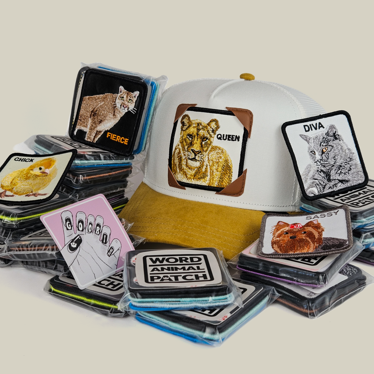 A white and tan Gal Pals Patch Pack with GB Patch Corner Trucker - Tan decorated with a lion design, surrounded by a stack of assorted animal patches in clear plastic sleeves.