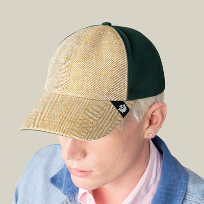 A person wearing the Goorin Bros. Zig Zag Tailored Baseball hat in green and beige herringbone, with a light blue jacket, pink checkered shirt, and stud earring, facing slightly downward.