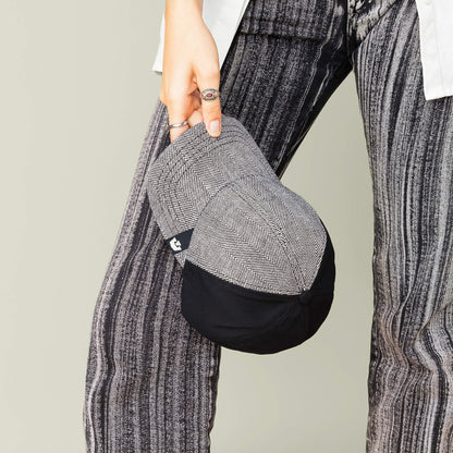 A person holds the Goorin Bros. Zig Zag Tailored Baseball hat, featuring a black and gray herringbone pattern, next to their striped pants; only the lower body and hand are visible.