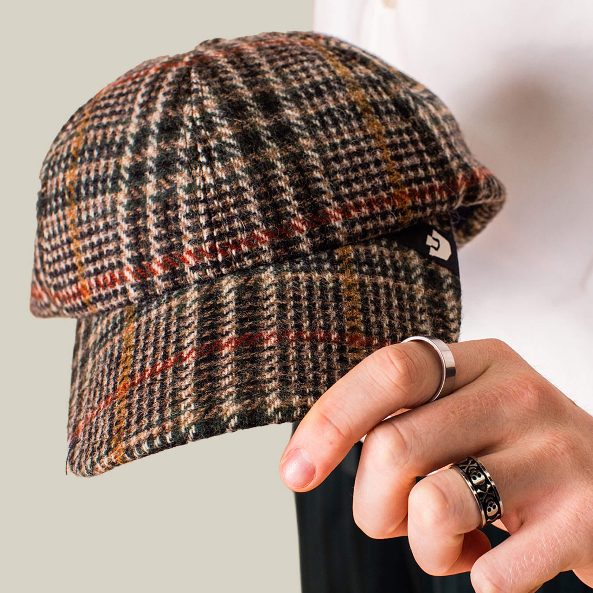 A hand with silver rings holds the Goorin Bros. Cozy Tailored Baseball cap, a classic six-panel design in plaid shades of brown, red, and green.