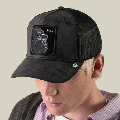 A person wearing the Goorin Bros. Velour Boss cap—a black velour trucker hat with breathable mesh and a patch featuring a gorilla and the word “BOSS”—looking downward.