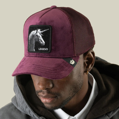 A man wearing the Goorin Bros. Velour Legend, a burgundy velour trucker hat with a breathable mesh back and a monochrome unicorn patch reading “Legend” on the front, looks slightly downward while dressed in a grey hoodie and white shirt.
