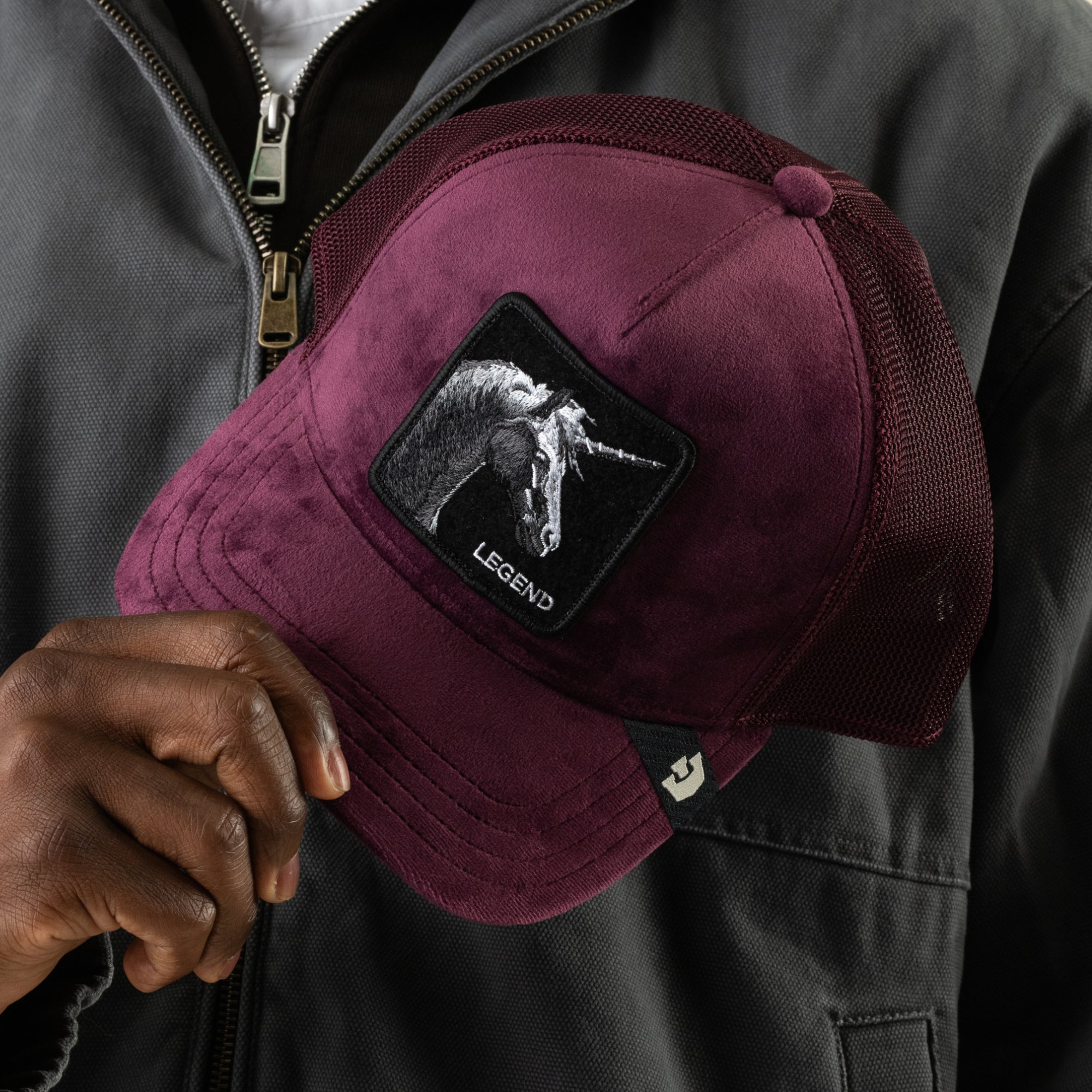 A person holds the Goorin Bros. Velour Legend trucker hat in burgundy, featuring a monochrome unicorn patch and a breathable mesh back, while wearing a dark jacket with a visible zipper.