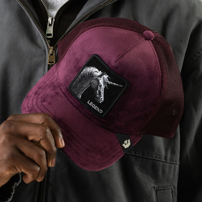 A person holds the Goorin Bros. Velour Legend trucker hat in burgundy, featuring a monochrome unicorn patch and a breathable mesh back, while wearing a dark jacket with a visible zipper.