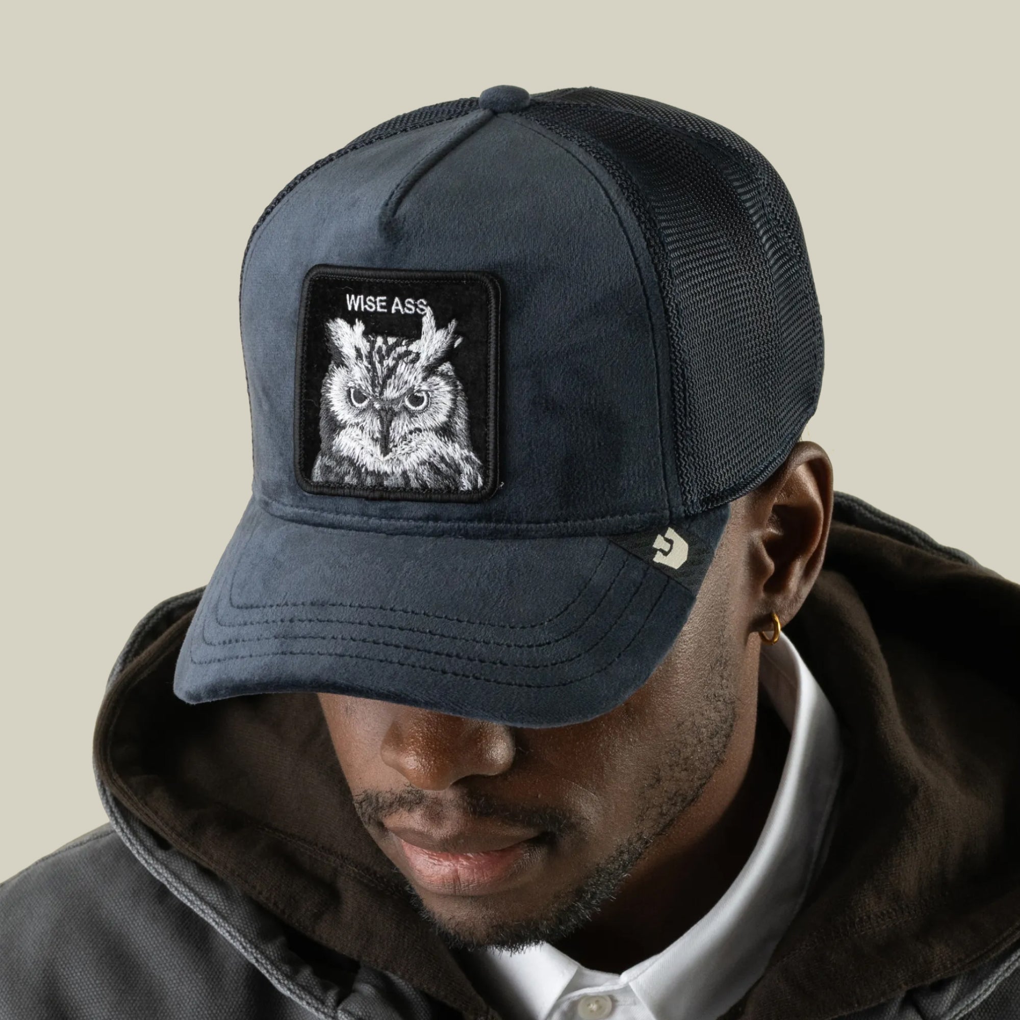 A man wearing a dark Goorin Bros. Velour Wise Ass trucker hat featuring an owl patch that reads WISE ASS on the front, dressed in a dark hoodie and looking down.