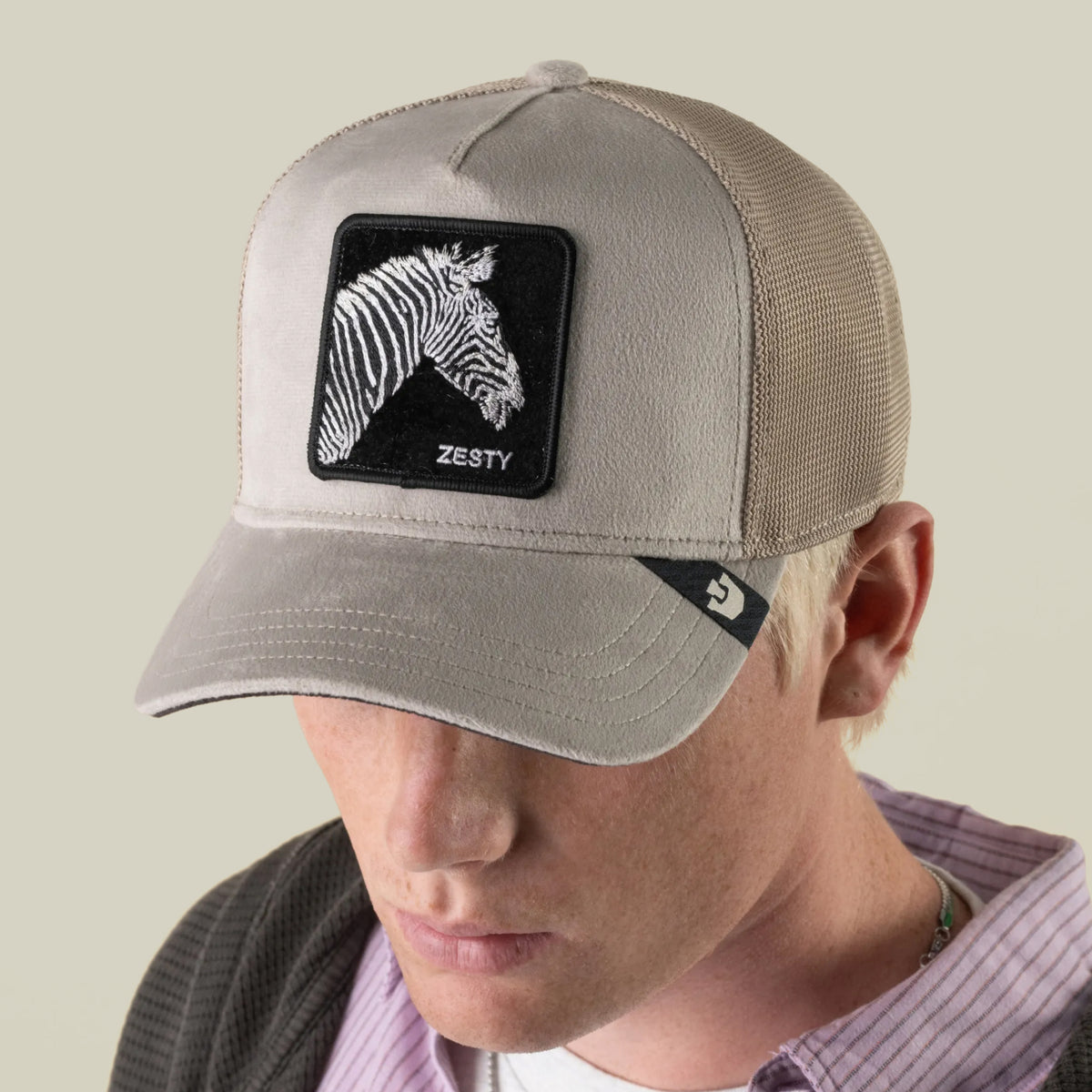 A person wearing the Goorin Bros. Velour Zesty beige mesh baseball cap, featuring a striking black-and-white zebra patch labeled “ZESTY” on the front panel, reminiscent of a classic monochrome zebra trucker hat.