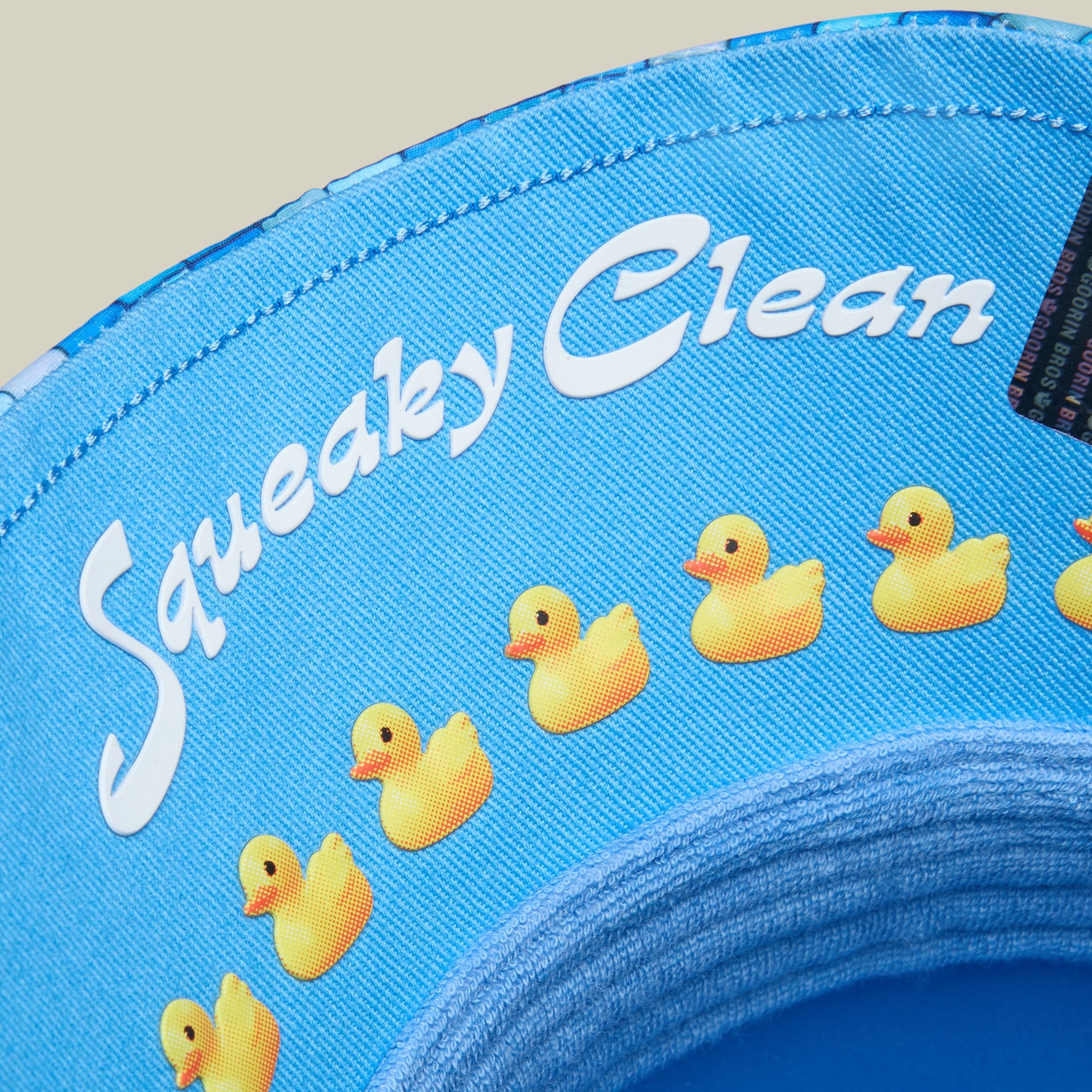 Close-up of a blue fabric item from Goorin Bros., featuring the "Squeaky Clean" text and a row of yellow rubber duck illustrations printed on it—perfect for adding some sudz fun to bath time.