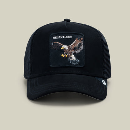 The Goorin Bros. Suede Relentless Eagle Trucker in Void features a black suede-feel brim, breathable mesh back, and a patch with an embroidered bald eagle topped by the word “RELENTLESS,” showcased against a plain light background.