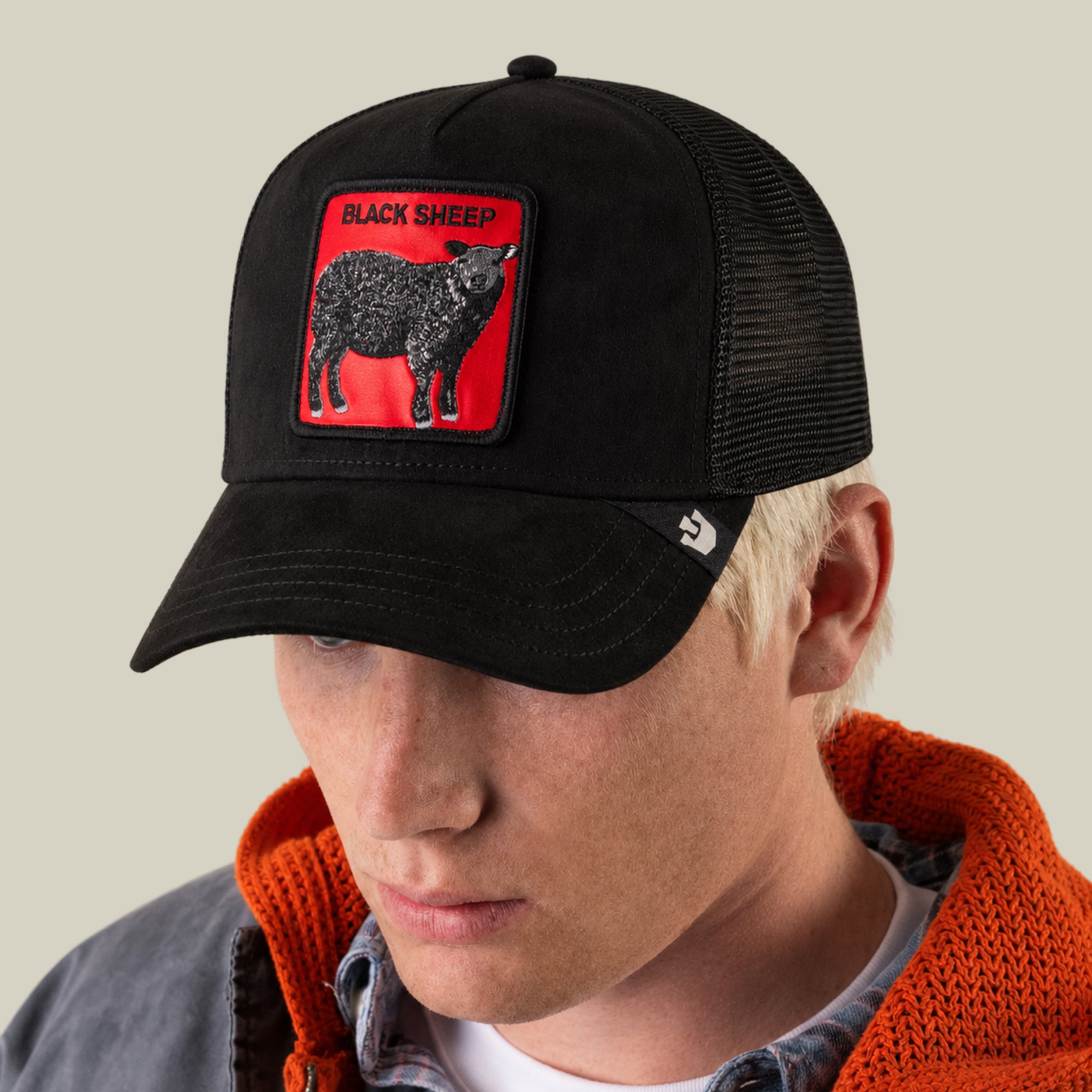A person with light hair is wearing the Goorin Bros. Pecora Nera Rossa trucker hat, which features a mesh back and a red patch displaying a black sheep and the text “BLACK SHEEP.” They are also dressed in a gray jacket with an orange hood.