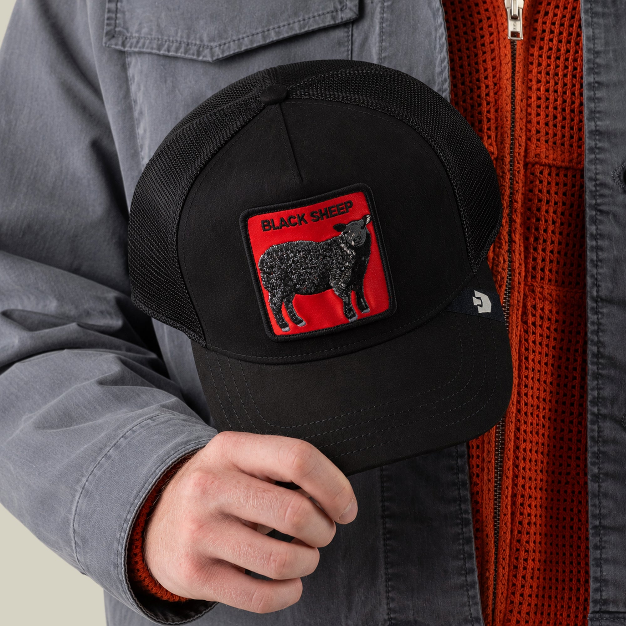 A person wearing a gray jacket and orange sweater holds the Goorin Bros. Pecora Nera Rossa trucker hat, featuring a mesh back and a patch with a black sheep and the words BLACK SHEEP on a red background, for a unique, casual style.