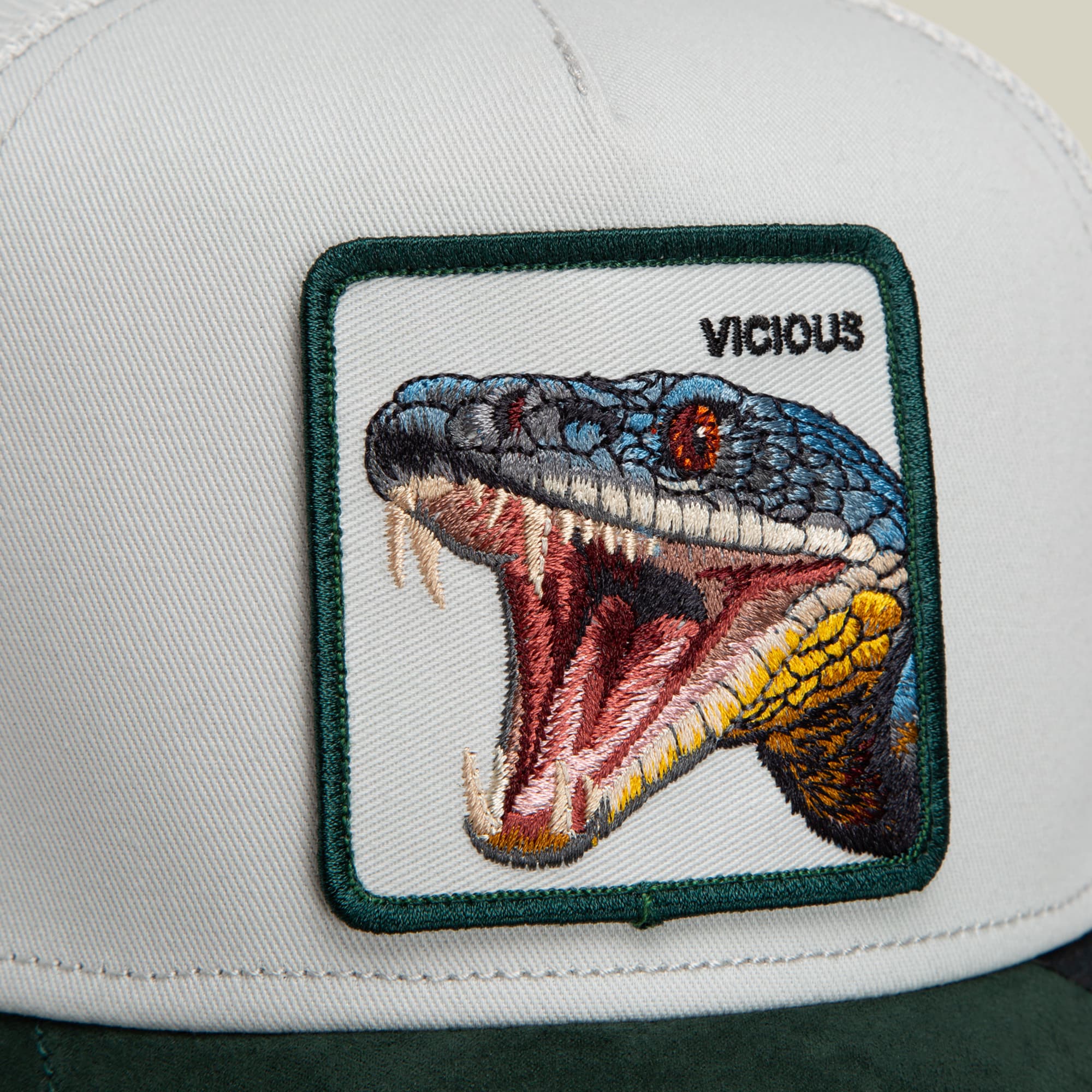 The Goorin Bros. Suede Snake trucker hat features a white front panel and green back with an embroidered patch of a vicious snake with its mouth open, detailed in blue, red, and yellow thread.