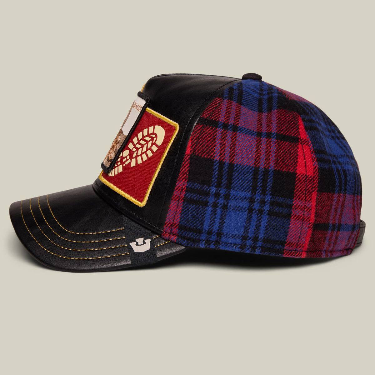 The Goorin Bros. Banger trucker hat features a black leather front with gold stitching, red and blue plaid wool back panel, and an embroidered red and gold leaf patch on the side.