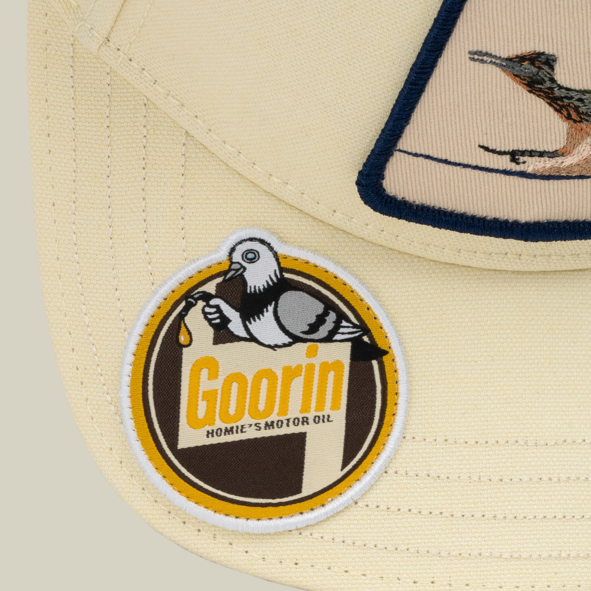 The Crude Guzzler by Goorin Bros is a cream and navy trucker hat featuring an embroidered vintage motor oil patch with a bald eagle design and yellow gold accents on the front panel.