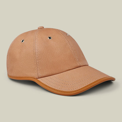 The Steiner by Goorin Bros. is a tan leather papa cap with stitched panels, metal eyelets, and a curved brim. It features an adjustable plaid strap and is shown against a light beige background.