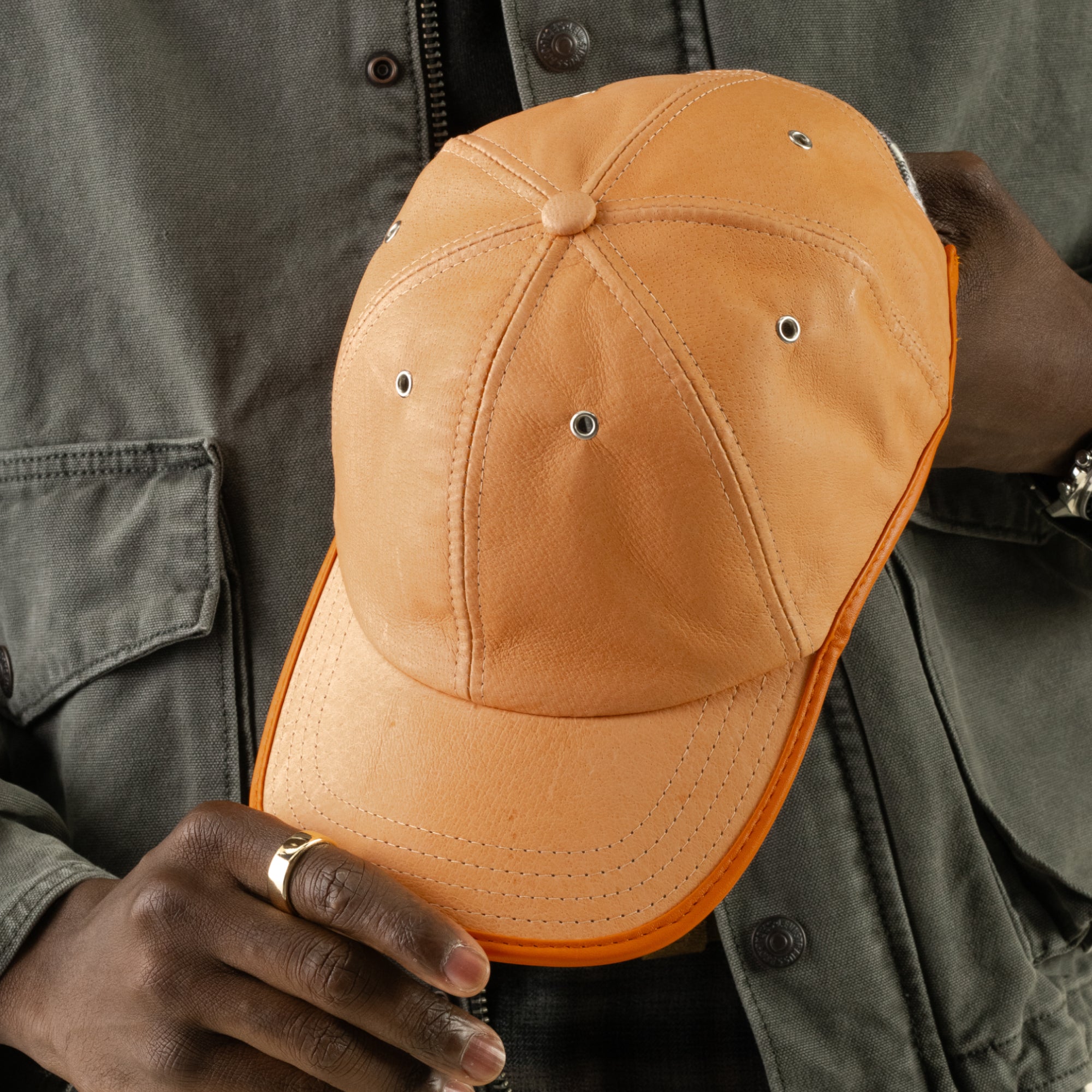 A person wearing a gray jacket holds the Steiner cap by Goorin Bros.—an orange suede cap with metal eyelets—displaying its front and top panels. The Steiner papa cap features an adjustable plaid strap at the back for a comfortable fit.