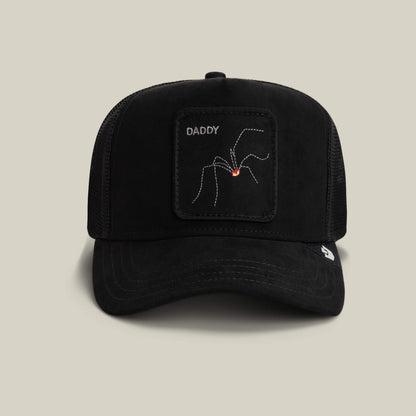 The Goorin Bros. Bold Suede Spider is a void black daddy trucker hat with a patch of an arachnid labeled DADDY and a small red mark, shown against a plain beige background.