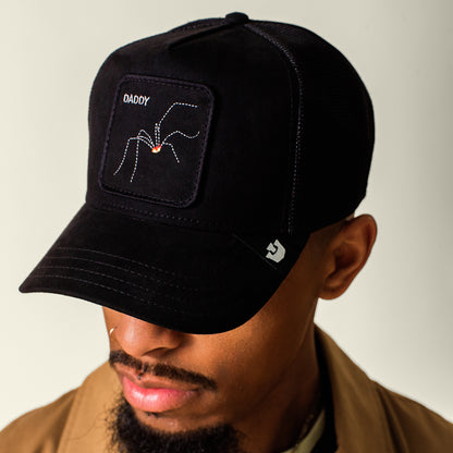 A man wears the Goorin Bros. Bold Suede Spider hat, which is black with an arachnid patch and "DADDY" embroidery. He also has a nose ring and is dressed in a brown microsuede jacket.