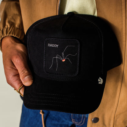 Someone holds a black Goorin Bros. Bold Suede Spider trucker cap featuring a stitched white spider with a red dot and the word DADDY on the front patch.