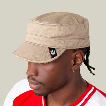 A person wears the "Home at Last" beige military-style cap by Goorin Bros., featuring a black and white patch on the side, paired with a red and white shirt against a plain light background.