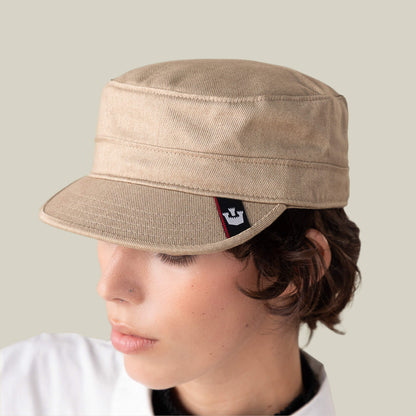 A person wearing the Goorin Bros. "Home at Last" beige military-style cap, featuring a small black and red logo tag, looks downward against a plain light background.