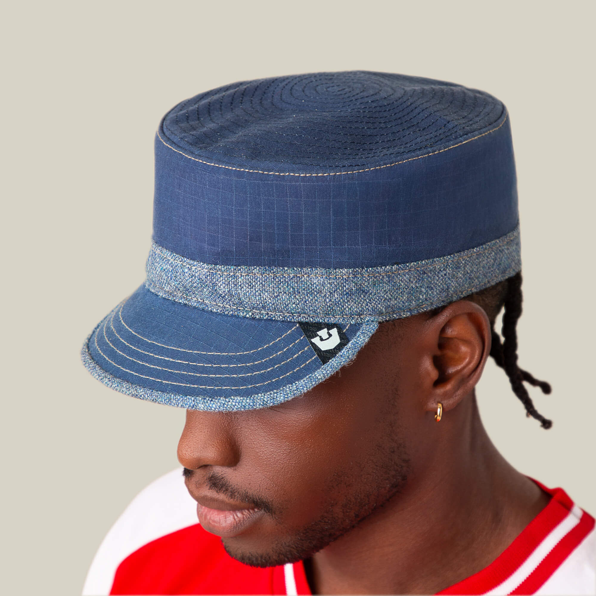 A person wears the Goorin Bros. Ampere, a blue flat-top cadet hat with a denim chambray brim, paired with a red and white shirt, against a plain light background.