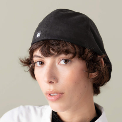 A person with short brown hair wears the Murray Way black flatcap by Goorin Bros. and a white 100% cotton top, looking at the camera against a neutral background.