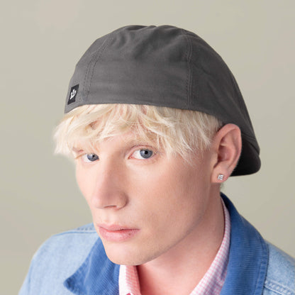 A person with platinum blonde hair wears the Goorin Bros. Murray Way gray flatcap backwards, a 100% cotton American-made denim jacket, stud earring, and pink checkered shirt, looking at the camera with a neutral expression.