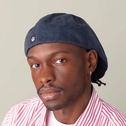 A man poses against a plain light background wearing the Goorin Bros. Murray Way blue flatcap, a gold hoop earring, and a red-and-white striped 100% cotton shirt.
