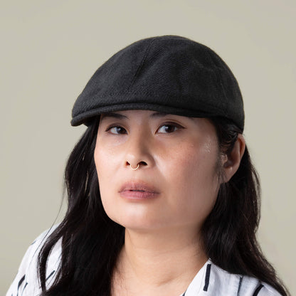 Person with long dark hair wears the Ronald Amos cap by Goorin Bros.—a black six-panel flat cap with a two-inch brim—and a white shirt with black stripes, looking directly at the camera with a neutral expression.