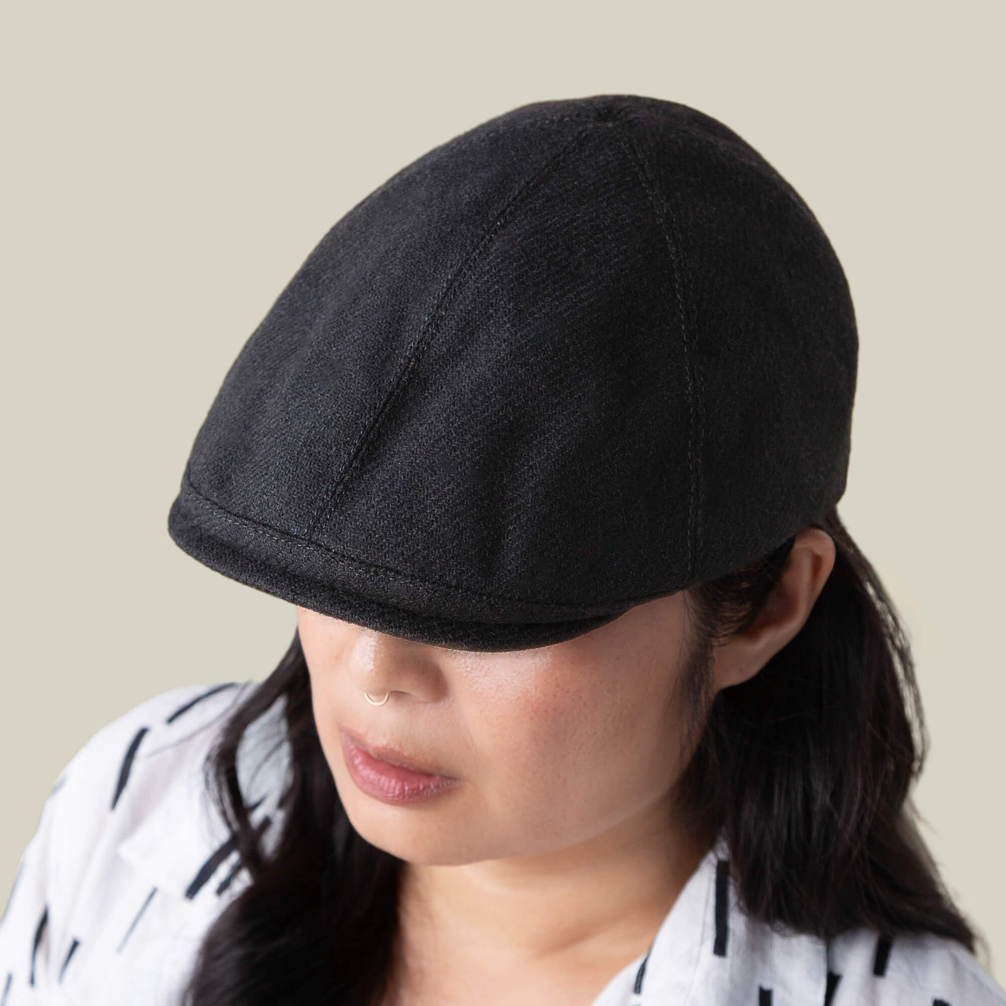 A person wearing the Goorin Bros. Ronald Amos black six-panel flat cap with a two-inch brim and a white patterned shirt, looking downward against a plain beige background.