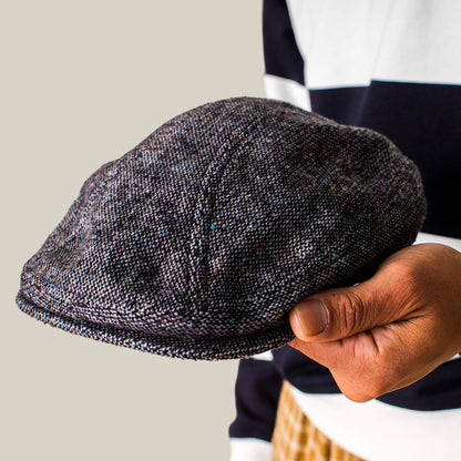 A person holds the Gibbins flatcap by Goorin Bros. in one hand, styled with a black-and-white striped long-sleeve shirt for a timeless men's fashion look.