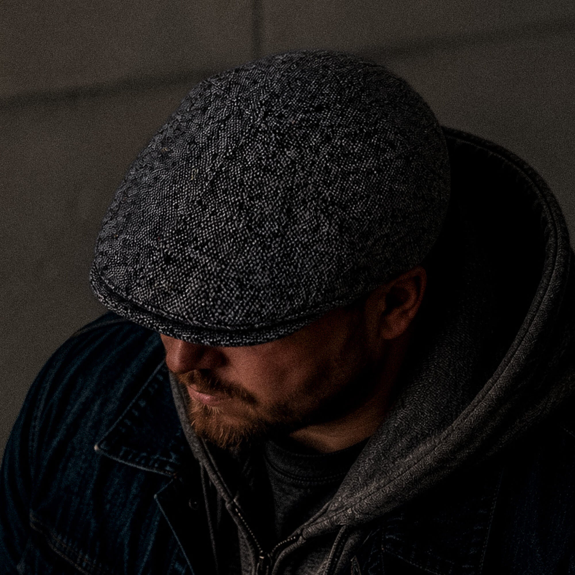 A man in the dim light wears the Goorin Bros. Newlands, a hand cut and sewn grey flecked flat cap, paired with a dark jacket and hoodie, looking downwards.