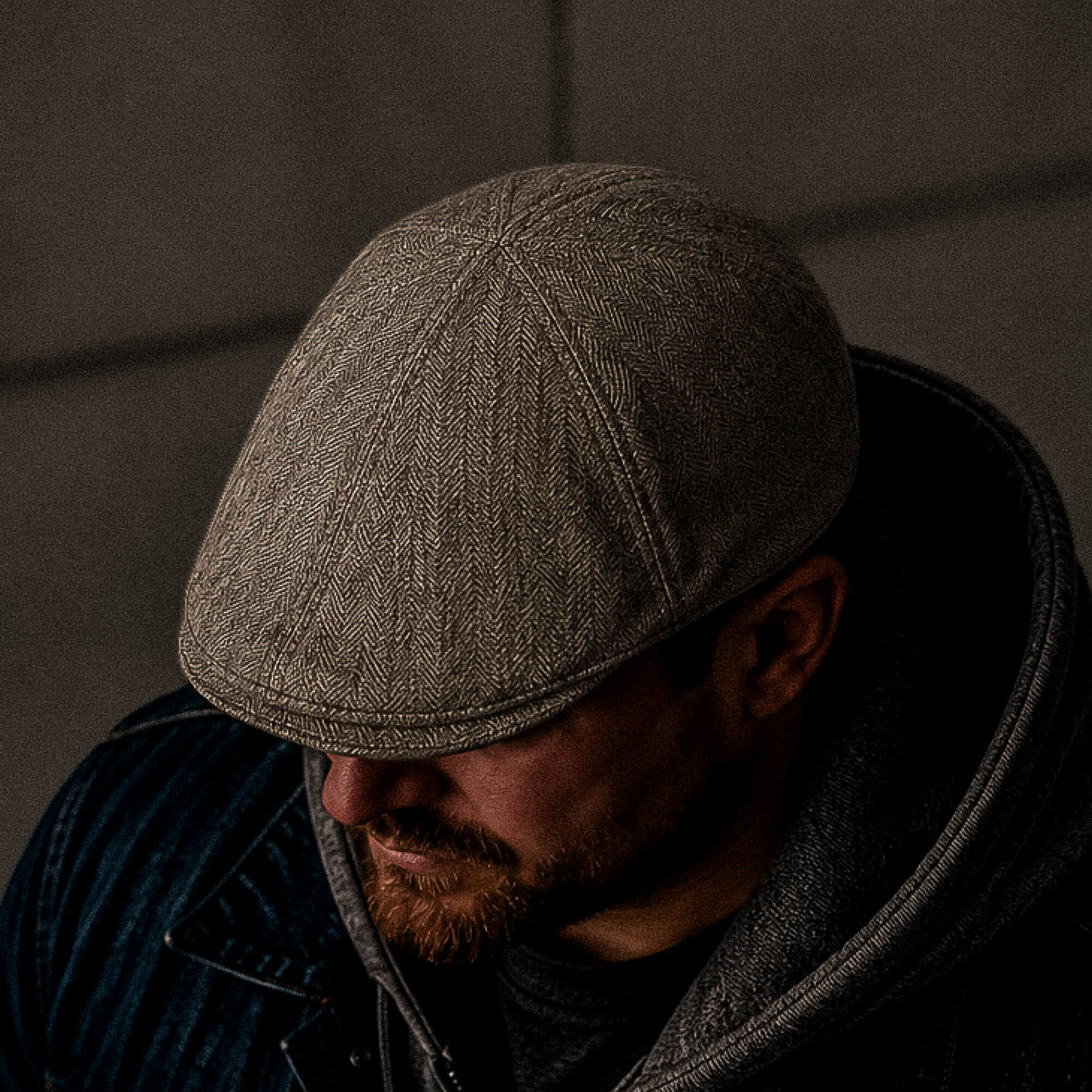 A man wearing the Goorin Bros. Harry Bones tan herringbone wool flatcap, paired with a hoodie and jacket, looks down in low lighting.