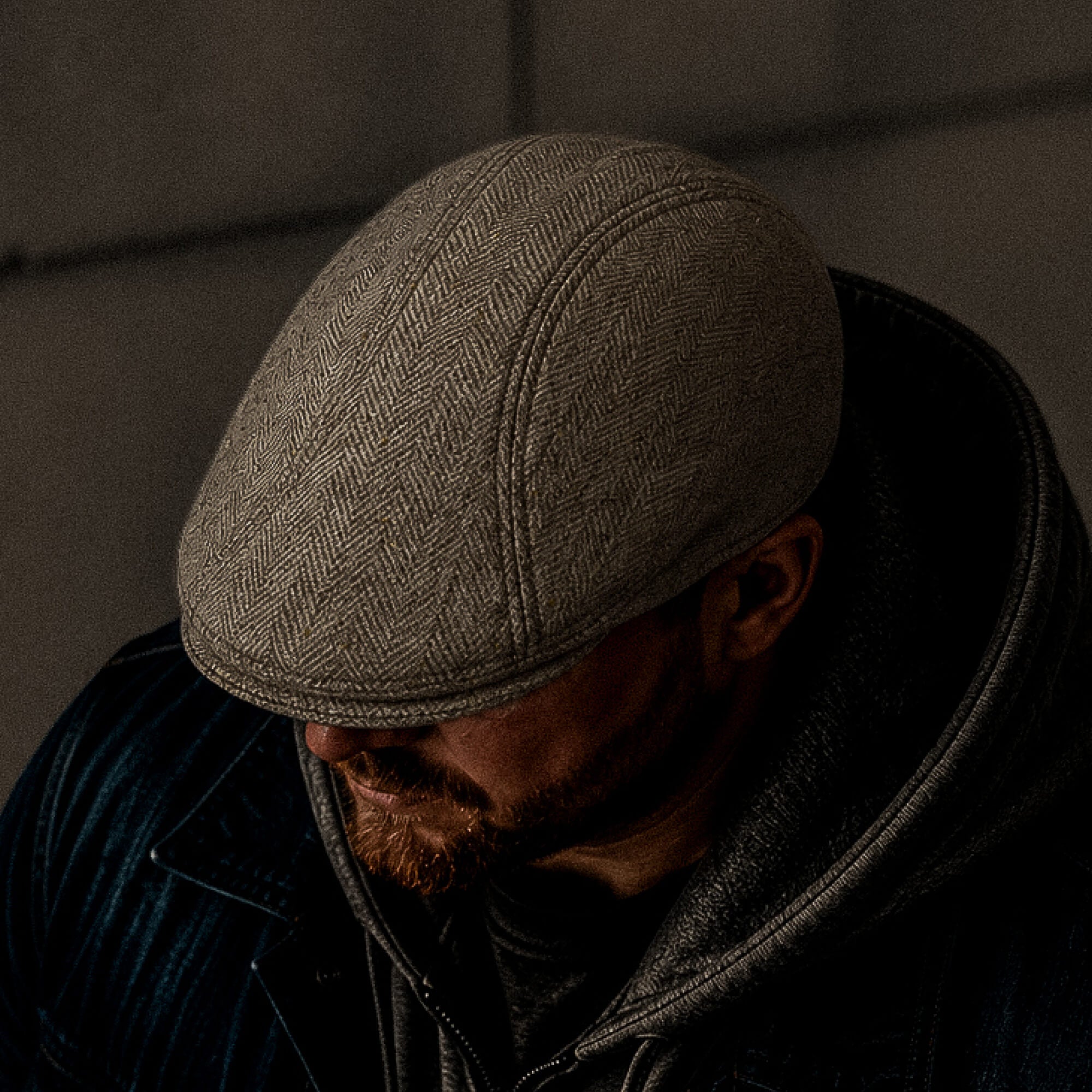 A bearded man wears the Iris Fields cream herringbone wool flatcap by Goorin Bros., paired with a hoodie and jacket. He looks down, his face partly in shadow. The handmade flatcap is crafted in the USA.