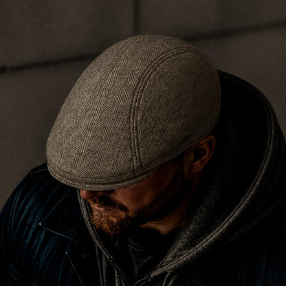 A bearded man wears the Iris Fields cream herringbone wool flatcap by Goorin Bros., paired with a hoodie and jacket. He looks down, his face partly in shadow. The handmade flatcap is crafted in the USA.