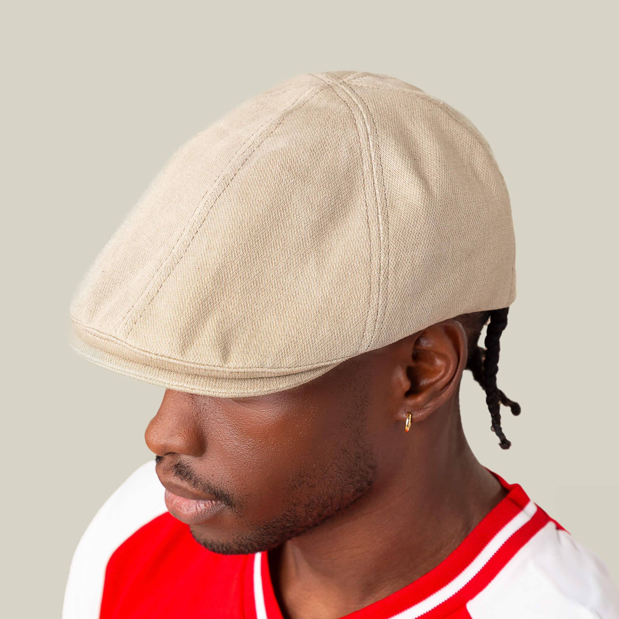 A man models the Goorin Bros. Noisy Benjamin Paul beige flatcap, paired with a red and white shirt and small hoop earring, set against a plain light background.