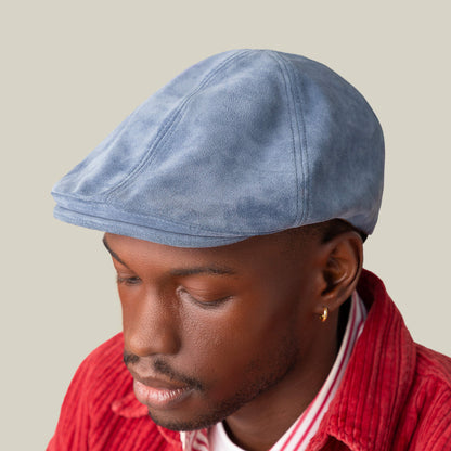 A man in the Cushy Benjamin Paul light blue suede flatcap by Goorin Bros., paired with a red corduroy jacket and striped shirt, looks downward against a plain background.