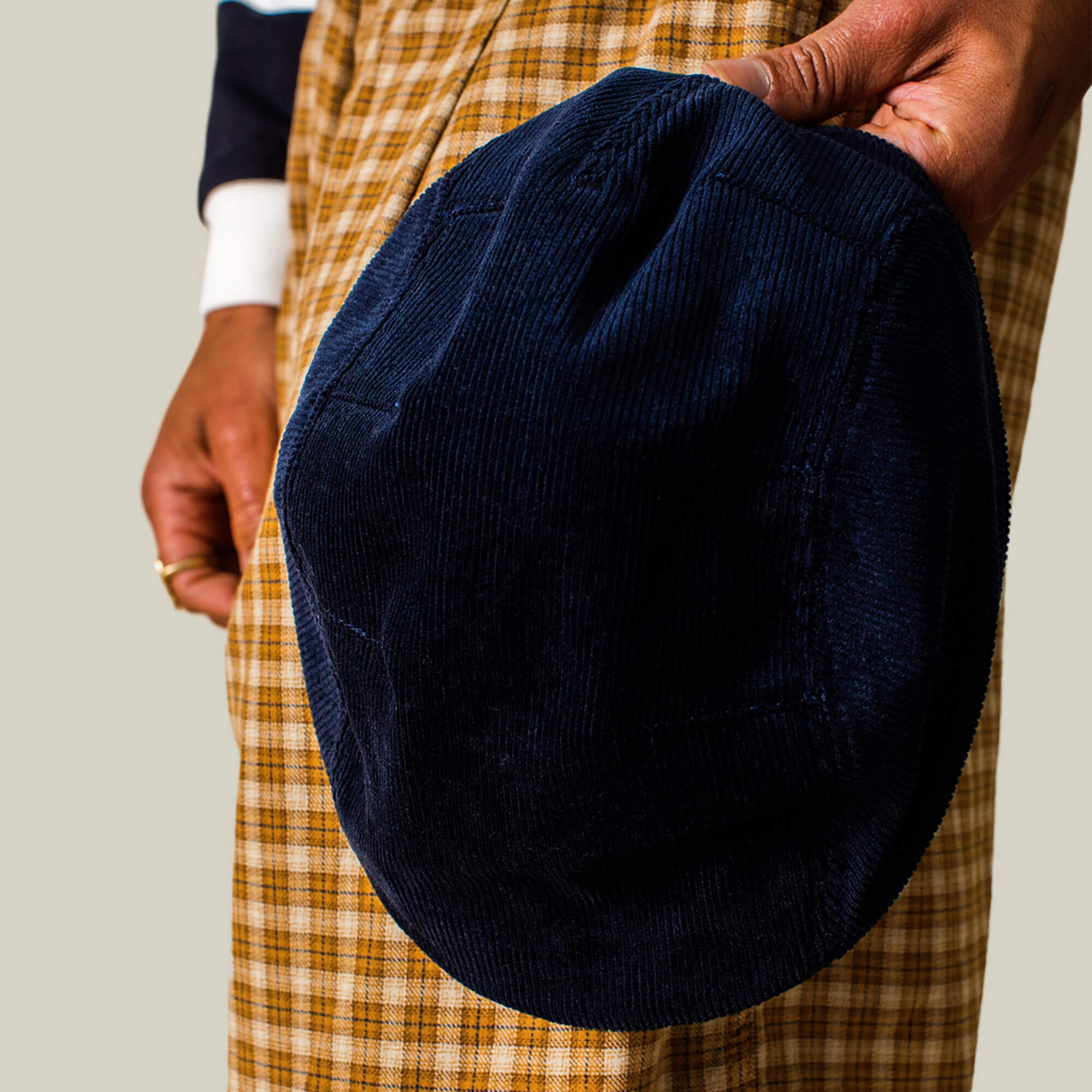 A person holding the Goorin Bros. Cozy Low Profile navy corduroy flatcap, dressed in a plaid garment and a white shirt with dark sleeves.