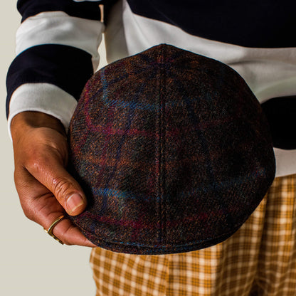 A person in a striped shirt and plaid pants holds the Cozy Benjamin Paul ivy cap by Goorin Bros. in one hand.