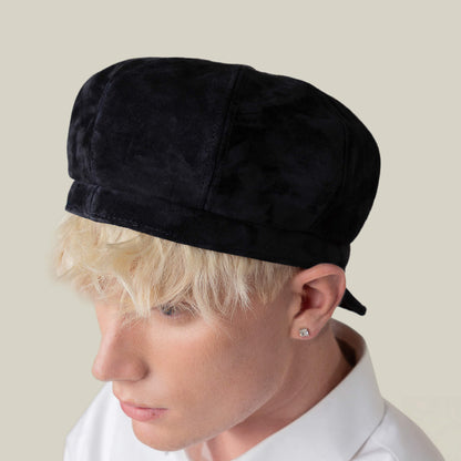 A person with short blond hair wears the "Ruthy" black cabbie flatcap by Goorin Bros. and a white collared shirt, shown from a side angle against a plain background.