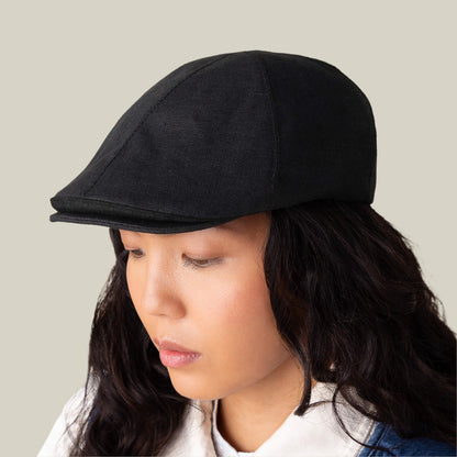 A person with long dark hair wears the Benjamin Paul three-season hat by Goorin Bros. with a white collared shirt, looking downward against a plain background.