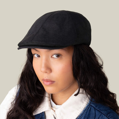 Someone with long dark hair models the Goorin Bros. Benjamin Paul, an everyday black linen hat, paired with a white collared shirt and blue vest, gazing directly at the camera against a plain background.
